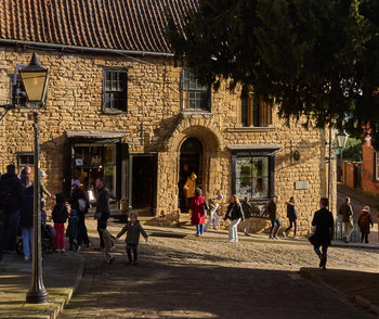 This urban photograph captures Lincoln’s famous Steep Hill in the heart of England, United Kingdom, during a bright winter afternoon. The image shows a charming stone building that houses Imperial Tea & Coffee, where people can be seen entering and leaving the shop, enjoying their winter shopping in this historic part of Lincoln. The photograph highlights the lively atmosphere as locals and visitors stroll along the cobbled street, framed by traditional English architecture. The wintry sunlight casts long shadows, adding to the inviting feel of the bustling street scene, and the presence of families, children, and individuals enjoying Steep Hill emphasizes its popularity as a social and cultural landmark in Lincoln, England.